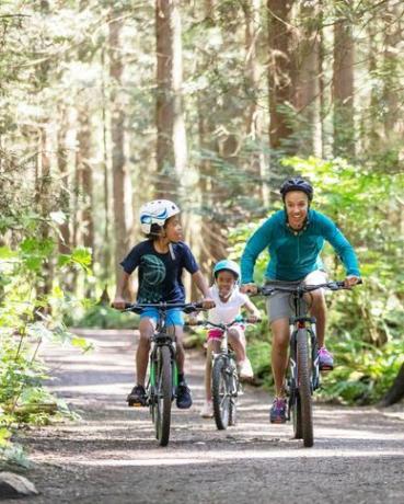 egy nő és két gyereke biciklizik az erdőben a napsütésben