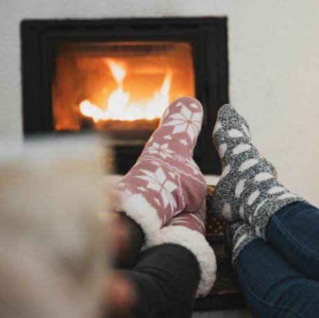 kilátás két barátnő mögül, akik a kandalló és a karácsonyfa mellett ülnek, lábukkal, téli zoknival a dohányzóasztalon, kávét vagy teát isznak