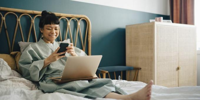 mosolygó nő laptop segítségével okos telefon az ágyon otthon