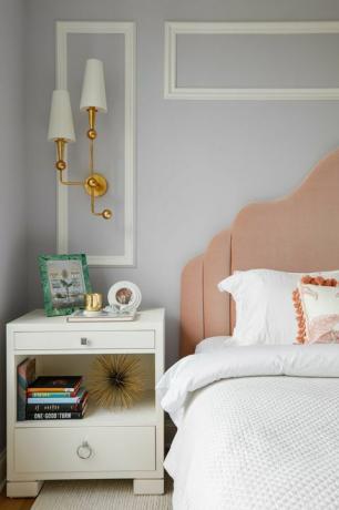 hálószoba, fehér linónusú ágy, fehér éjjeliszekrény, könyvek, rózsaszín fejtámla, fehér és arany burkolatok, fehér korona díszítés világosszürke kékre festett falakon